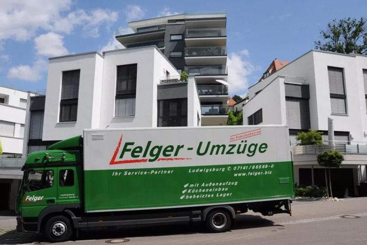 Ein grüner Umzugswagen mit der Aufschrift Felger-Umzüge  parkt vor modernen weißen Wohnhäusern vor einem blauen Himmel.